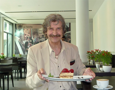 Filmfotograf Mike Gallus steht lächelnd im Ristorante Gennaro München-Sendling, einen Teller mit Tiramisu von Chefkoch Gennaro Donato in den Händen haltend neben einer auf dem Stehtisch stehenden Tasse Cafe