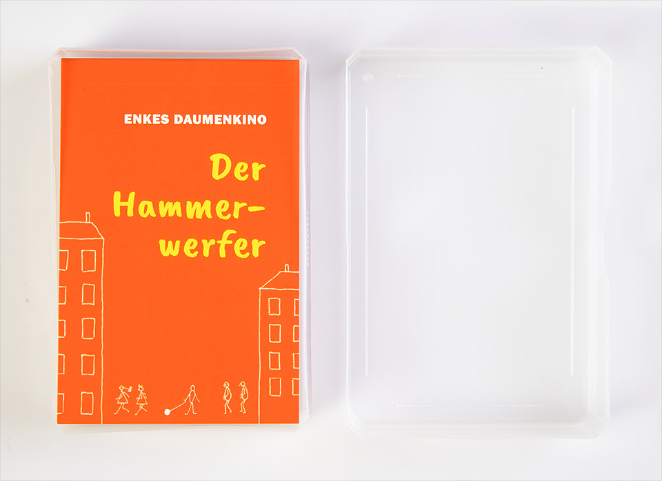 Leuchtend orange Vorderseite von Enkes Daumenkino "Der Hammerwerfer", das in einer geöffneten, durchsichtigen Kunststoff-Spielkartenschachtel mit daneben liegendem Deckel